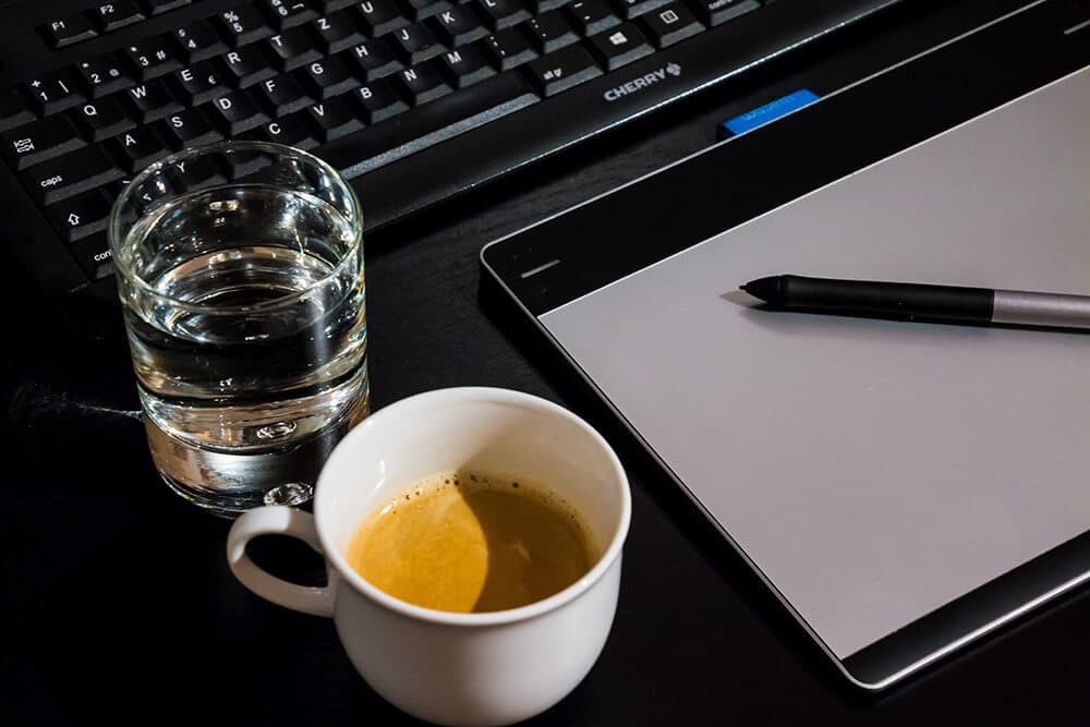 cup of coffee and water on desk next to pen and tablet and laptop